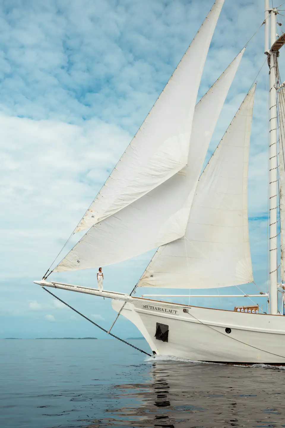 Mutiara Laut sailing yacht passing through a coastal Indonesian village landscape.